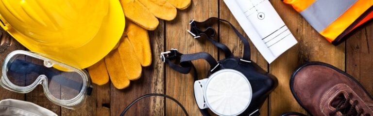 Équipements de sécurité pour le BTP : casque, gants, gilet fluorescent, chaussures et plans de chantier disposés sur une table en bois.